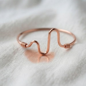 May include: A copper-coloured metal bangle bracelet featuring a central heartbeat-style wave pattern. The design includes two small sections of coiled wire detailing on either side of the central wave, creating a continuous, smooth metallic loop.