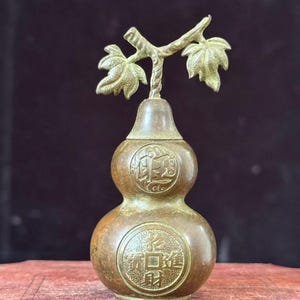 May include: A bronze-colored gourd-shaped decorative item with a branch and leaf design on top. The gourd has two sections and features circular embossed patterns with Chinese characters. The item is likely a decorative piece or a small container.