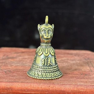 Puede incluir: Una pequeña campana de color bronce con una cara de mono en la parte superior. La campana tiene un lazo para colgar y presenta patrones decorativos y caracteres chinos. La campana está sobre una superficie de madera.