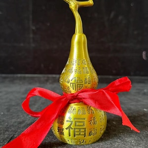 May include: A gold-colored, gourd-shaped ornament featuring a stem at the top and numerous embossed Chinese characters, including the prominent character for fortune, wrapped with a bright red satin ribbon tied in a bow around its center.