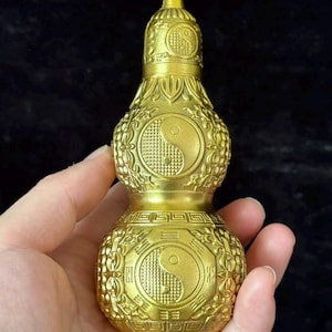 May include: A gold-coloured metal gourd-shaped vessel featuring embossed yin-yang symbols on both the upper and lower sections. The surface is decorated with intricate relief patterns, including geometric borders and stylized foliage motifs.