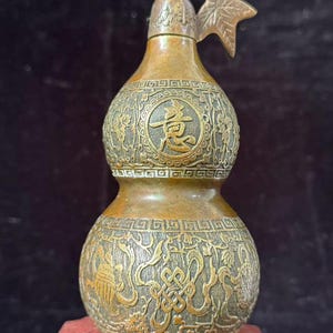 May include: A bronze-toned metal gourd-shaped vessel featuring intricate embossed patterns, including a central circular medallion with the Chinese character 意, a decorative stem with a leaf, and geometric border designs around the body.