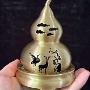 May include: A brass-colored metal incense burner shaped like a gourd, featuring cut-out designs of a crane, a deer, and a tree on the body, with cloud-shaped cut-outs near the top, resting on a matching circular base with decorative edges.