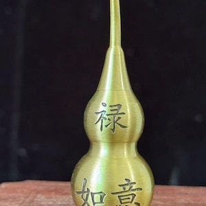 May include: A brass-colored, gourd-shaped decorative item with a slender neck and a bulbous base. The item features Chinese characters etched into its surface. The overall design is symmetrical and stands upright, showcasing a traditional aesthetic.