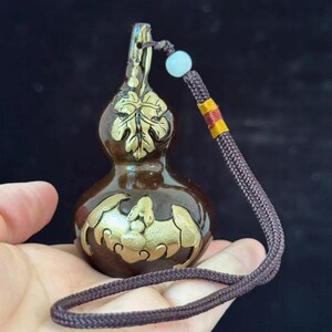 May include: A dark brown, gourd-shaped pendant featuring gold-colored embossed details of a leaf on the upper section and a bat on the lower section. A brown braided cord is attached to the top, accented with a small light blue bead.