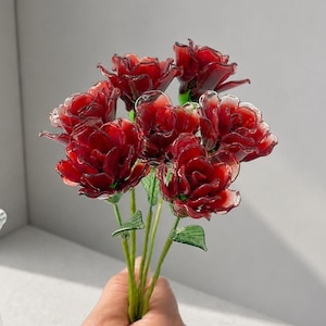 May include: A bouquet of seven deep red glass roses with translucent petals and green glass stems and leaves. Each rose features layered, curved petals that catch the light, creating a glossy, reflective appearance throughout the floral arrangement.