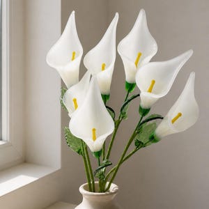 May include: A bouquet of six handcrafted glass calla lilies with translucent white petals and bright yellow spadices. Each flower features a green glass stem and textured green leaves, all arranged in a small, neutral-toned ceramic vase.