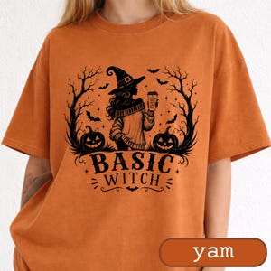 May include: A burnt orange short-sleeve t-shirt featuring a black graphic print of a witch wearing a hat, holding a cup labelled Pumpkin Spice Latte, surrounded by bare trees, bats, and two jack-o'-lanterns with the text Basic Witch below.