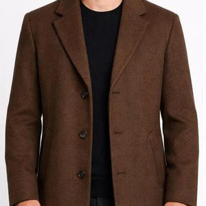 May include: A brown overcoat with a notched lapel and three-button closure. The coat has two front pockets and is worn over a black sweater. The coat is made of a wool-like material.