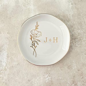 May include: A round white ceramic trinket dish with an irregular edge and a gold-colored rim. The center features a gold-colored line art illustration of a single flower with leaves and the text J + H printed in a matching gold-colored font.