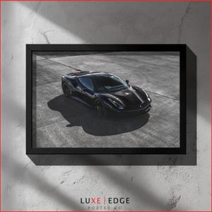 Peut inclure: Impression encadrée d'une voiture de sport noire sur une surface en béton. La finition brillante de la voiture reflète la lumière, et l'image est placée contre un mur texturé blanc cassé. L'impression est encadrée d'un bord noir, avec le logo Luxe Edge Poster Co. en bas.