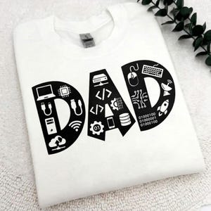 Peut inclure: T-shirt blanc avec le mot "DAD" en noir. Le design est composé d'icônes technologiques telles qu'un ordinateur portable, une souris, un clavier et du code binaire. Un cadeau idéal pour un père féru de technologie.