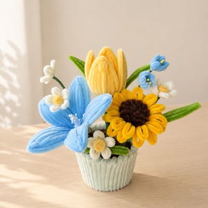 May include: A handmade floral arrangement featuring a yellow tulip, a blue lily, and a yellow sunflower with a brown center. The flowers are arranged in a light green, ribbed container. Green leaves and small white flowers complete the display.