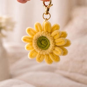 May include: A yellow and green sunflower keychain with a gold-colored clasp. The flower has soft, textured petals and a green center. The keychain is held up against a soft, blurred background.
