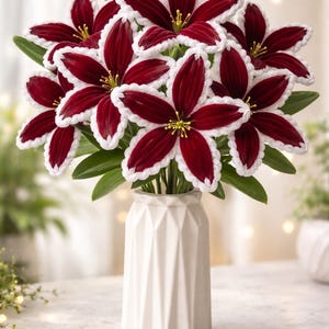 May include: A vase of deep red flowers with white edging and yellow centres. The flowers are in a white geometric vase. The background is blurred. The flowers have green leaves.