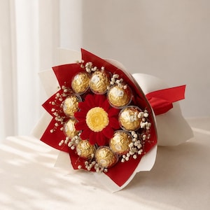 May include: A bouquet of golden chocolate candies arranged around a red flower. The bouquet is wrapped in red and white paper and tied with a red ribbon. Small white flowers are interspersed throughout. The candies are individually wrapped.
