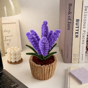 May include: A handcrafted, purple, fuzzy flower arrangement in a tan, ribbed pot. The faux plant has green leaves and sits on a white desk next to books, a laptop, a candle, and a notepad.