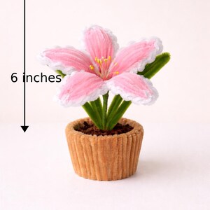 May include: A decorative artificial flower in a small pot. The pink flower has white edges and yellow stamens. The pot is brown and has a ribbed texture. The flower is approximately 6 inches tall and 5 inches wide.