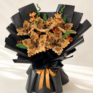 May include: A bouquet of handcrafted flowers made from textured tan and brown leopard-print fabric, accented with small orange blossoms and green stems. The arrangement is wrapped in black paper and tied with a gold ribbon at the base.
