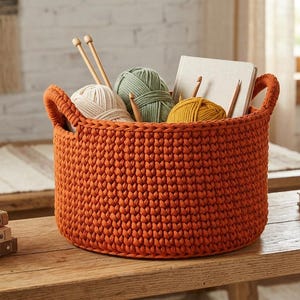 May include: A round, burnt orange crocheted storage basket with two integrated handles. The basket is filled with three balls of yarn in cream, sage green, and mustard yellow, along with wooden knitting needles and a light-colored notebook.