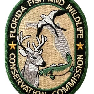 Puede incluir: Parche bordado con el logotipo de la Comisión de Conservación de la Pesca y la Vida Silvestre de Florida. El diseño incluye un ciervo, un pez y un pájaro, con texto alrededor del borde. El parche es verde, beige y blanco.