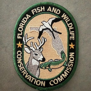 Puede incluir: Parche bordado con la inscripción "FLORIDA FISH AND WILDLIFE CONSERVATION COMMISSION". El parche muestra un ciervo, un pez y aves, sobre un fondo beige con un borde verde, ideal para los amantes de la naturaleza.