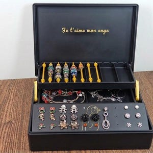 Puede incluir: Caja de joyería negra con múltiples compartimentos, llena de varios aretes, pulseras y collares. La caja tiene una tapa con bisagras con el texto "Je t'aime mon ange" en letras doradas. La caja está abierta, revelando las joyas en su interior.