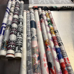 May include: A collection of rolled-up printed materials with various designs and text, including the phone number 601-249-5725. The rolls feature floral patterns, nautical themes, and graphic text, with a machine in the background.