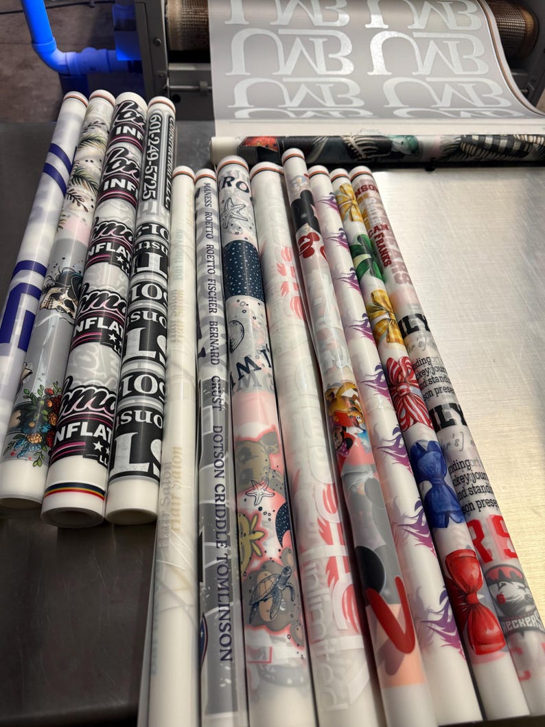 May include: A collection of rolled-up printed materials with various designs and text, including the phone number 601-249-5725. The rolls feature floral patterns, nautical themes, and graphic text, with a machine in the background.
