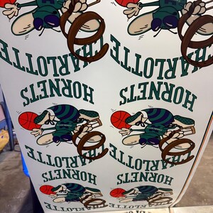 May include: A sheet of paper printed with a repeating pattern featuring the Charlotte Hornets logo. The logo includes a cartoonish bee holding a basketball, with the team name and city name in green text.
