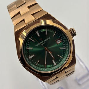 May include: A rose gold wristwatch with a green dial. The watch features a date window at the 3 o'clock position and a textured band. The watch face has the text "VACHERON CONSTANTIN GENEVE".