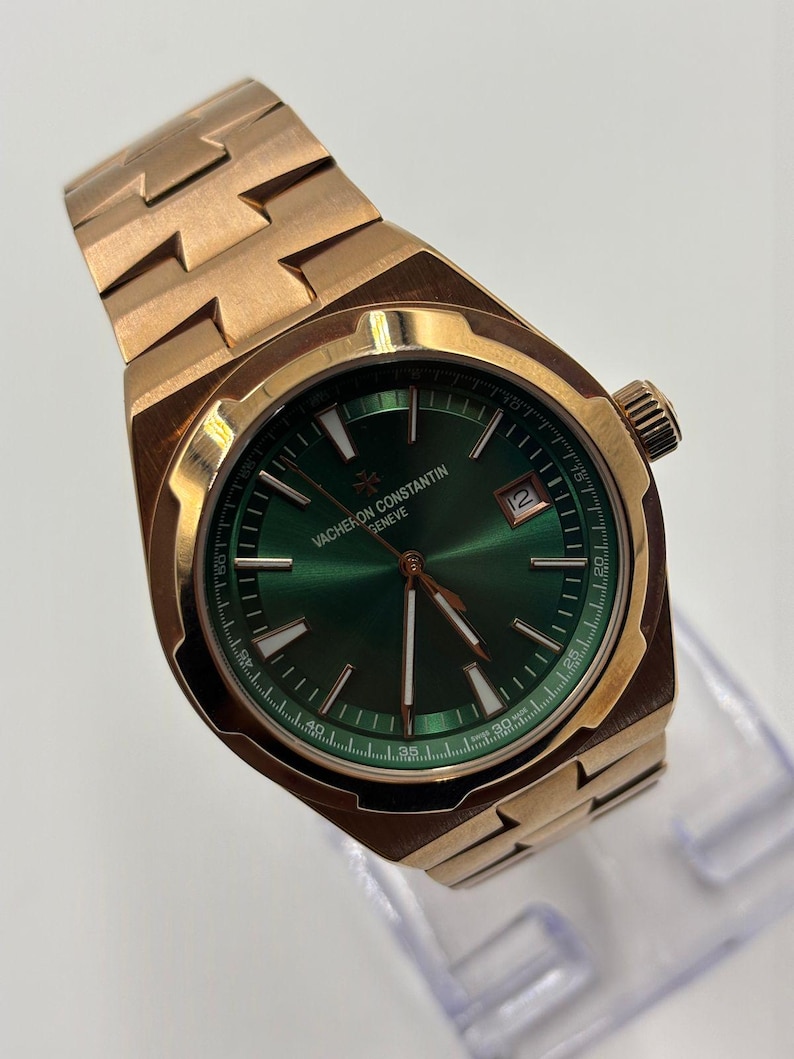 May include: A rose gold wristwatch with a green dial. The watch features a date window at the 3 o'clock position and a textured band. The watch face has the text "VACHERON CONSTANTIN GENEVE".