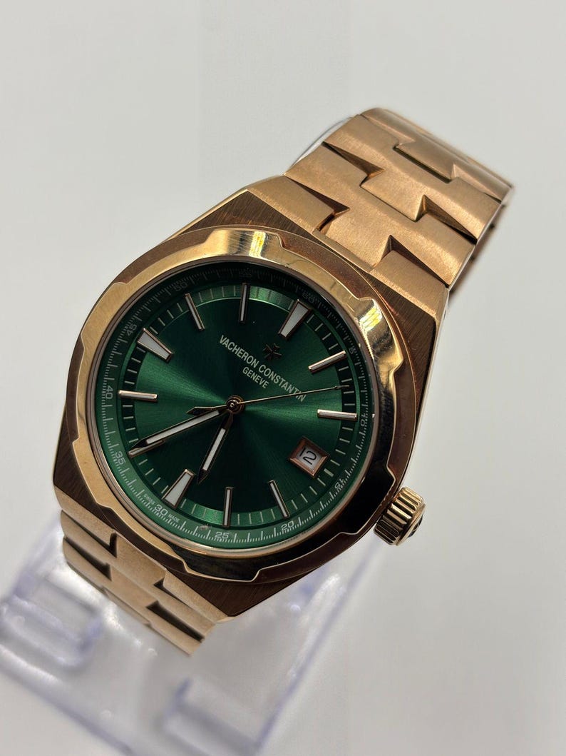 May include: A gold-toned wristwatch with a green face. The watch features a date window and a gold-toned band. The watch face has gold-toned hands and markers. The watch is displayed on a clear stand.