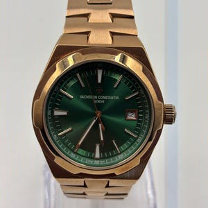 May include: A gold-toned Vacheron Constantin watch with a green dial. The watch features a date window at the 3 o'clock position and a gold-toned bracelet. The watch is displayed on a clear stand.