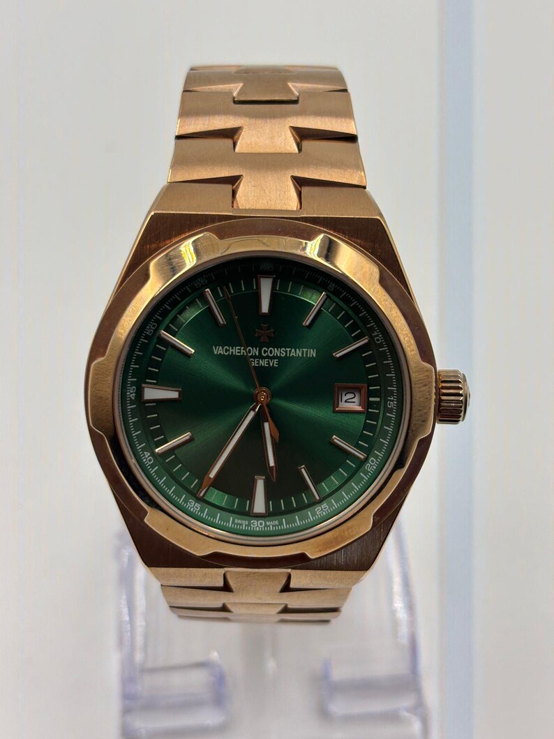 May include: A gold-toned Vacheron Constantin watch with a green dial. The watch features a date window at the 3 o'clock position and a gold-toned bracelet. The watch is displayed on a clear stand.