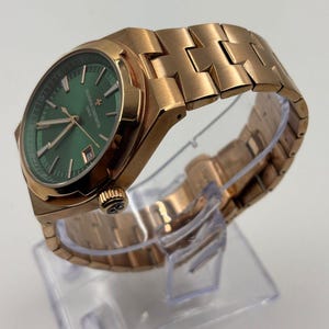 May include: A rose gold wristwatch with a green dial. The watch features a metallic band and a date display. The watch is displayed on a clear plastic stand. The watch face has the text "VACHERON CONSTANTIN GENEVE".