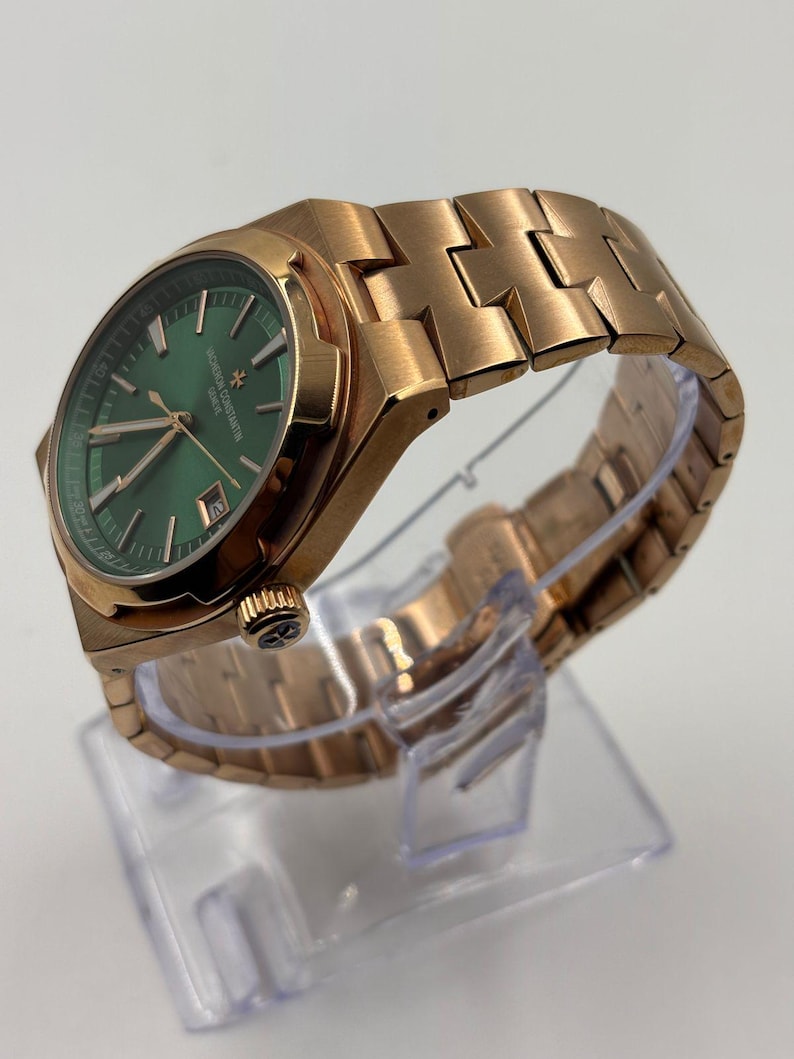 May include: A rose gold wristwatch with a green dial. The watch features a metallic band and a date display. The watch is displayed on a clear plastic stand. The watch face has the text "VACHERON CONSTANTIN GENEVE".