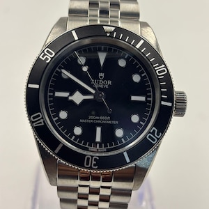 Puede incluir: Un reloj de pulsera de acero inoxidable con esfera negra y bisel giratorio negro. El reloj presenta marcadores de hora y agujas blancos, y el texto "TUDOR GENEVE" es visible en la esfera. La correa del reloj es de acero inoxidable.