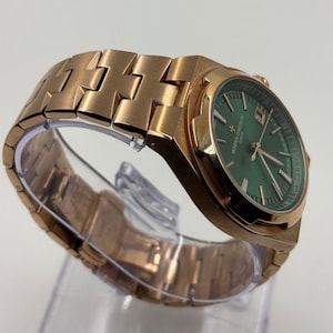 May include: A rose gold wristwatch with a green dial. The watch features a metallic band and a date display. The watch is displayed on a clear plastic stand. The watch face has the text "VACHERON CONSTANTIN GENEVE".