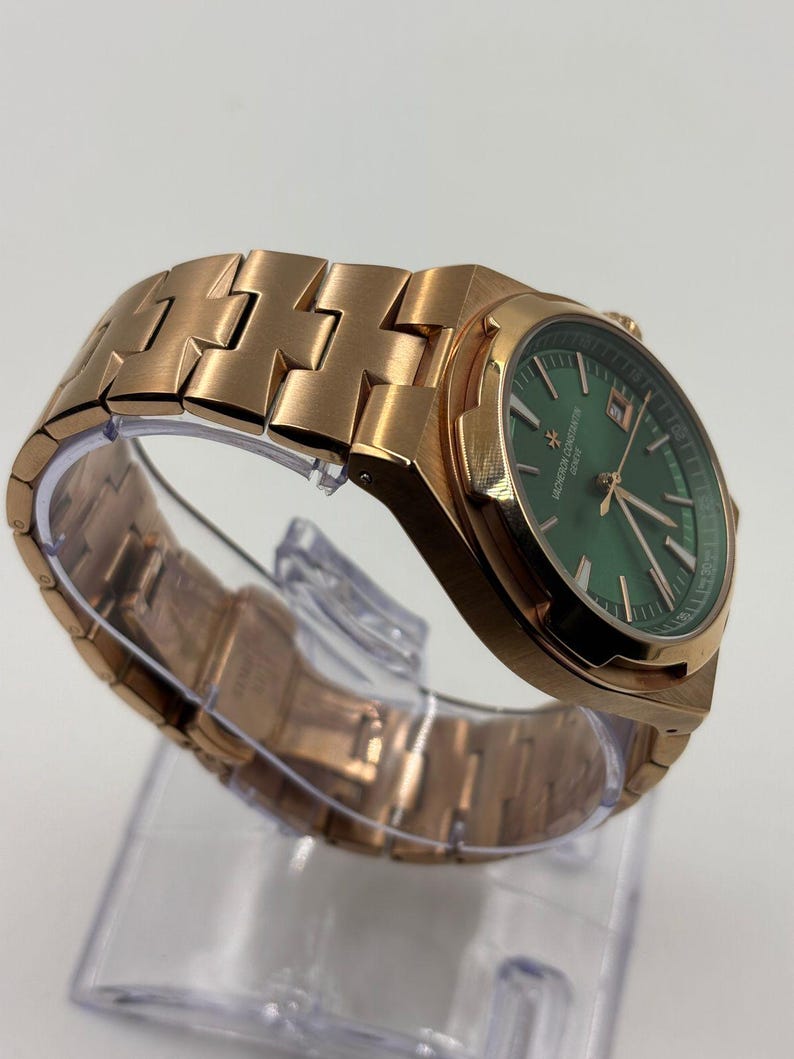 May include: A rose gold wristwatch with a green dial. The watch features a metallic band and a date display. The watch is displayed on a clear plastic stand. The watch face has the text "VACHERON CONSTANTIN GENEVE".
