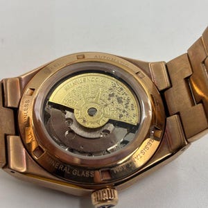 May include: Close-up of a rose gold-toned automatic wristwatch. The watch features a transparent back, revealing the intricate inner workings and a gold-colored rotor. The watch band and case are a matching rose gold color, with the words "STEEL", "AUTOMATIC", "ALL STAINLESS", "MINERAL GLASS", and "RESISTANT 50M" visible.