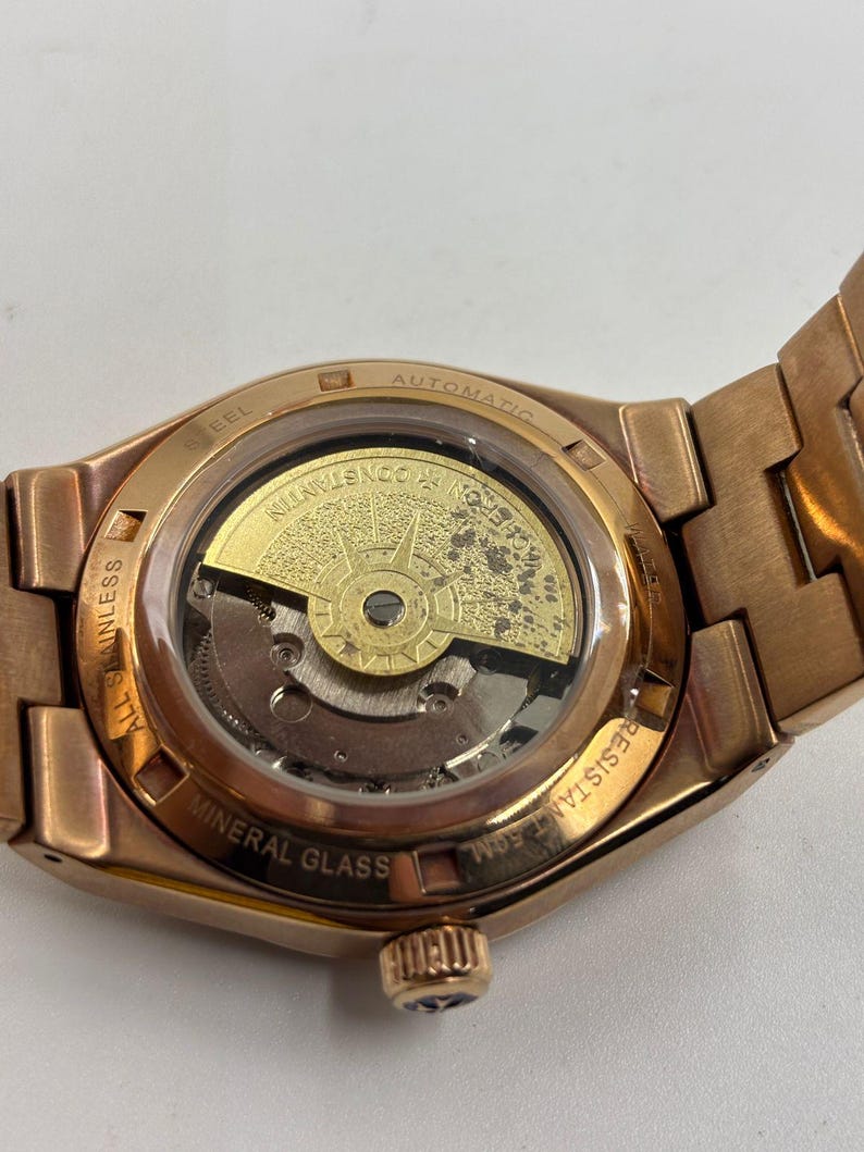 May include: Close-up of a rose gold-toned automatic wristwatch. The watch features a transparent back, revealing the intricate inner workings and a gold-colored rotor. The watch band and case are a matching rose gold color, with the words "STEEL", "AUTOMATIC", "ALL STAINLESS", "MINERAL GLASS", and "RESISTANT 50M" visible.