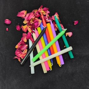 Puede incluir: Una colección de velas finas y cónicas de varios colores, incluyendo negro, blanco, rojo, naranja, amarillo, verde, azul, morado y rosa, dispuestas junto a pétalos de rosa secos de color rosa y amarillo sobre una superficie oscura.