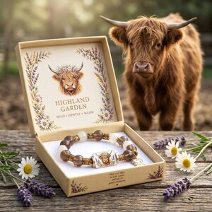 May include: A beaded bracelet with translucent brown and white beads and a rectangular charm featuring a Highland cow illustration, presented in a cardboard box labelled Highland Garden with floral accents and a Highland cow graphic.