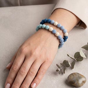May include: Two stretch bracelets made of porous, round lava stone beads in a gradient of colours. The beads transition from dark navy blue to medium blue, light blue, and off-white, arranged in a repeating pattern on an elastic cord.
