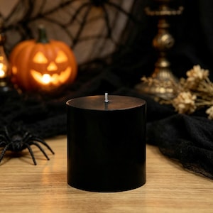May include: A solid black cylindrical pillar candle with a single white wick centered on the flat top surface. The candle has a smooth, matte finish and stands upright, presenting a simple, uniform design without any additional patterns or textures.