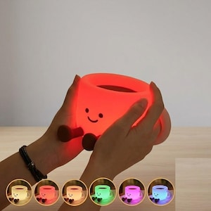 May include: A glowing, red, cup-shaped night light with a simple, printed smiling face and two small, dark brown, dangling legs. The light is shown in a variety of colors including white, red, yellow, green, pink, and blue in the small circular insets.