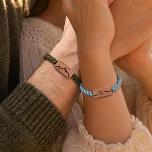 Puede incluir: Dos pulseras de cordón trenzado con dijes rectangulares de madera. Una pulsera tiene un cordón verde oliva oscuro con un grabado de montañas, mientras que la otra tiene un cordón azul claro con un grabado de una ola en el dije de madera.