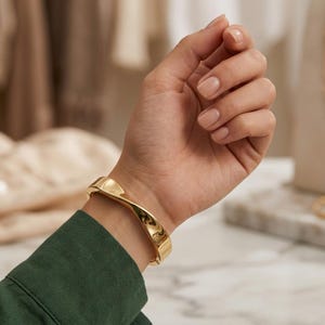 May include: A polished gold-tone hinged bangle bracelet featuring a smooth, twisted, and contoured design. The bracelet has a reflective metallic surface and a secure clasp mechanism, worn on the wrist above a dark green long-sleeved garment.