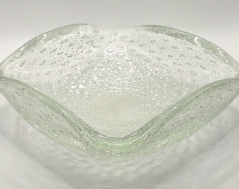 Könnte beinhalten: Eine klare Glasschale mit quadratischer Form und abgerundeten Ecken, die eine texturierte Oberfläche mit zahlreichen kleinen, eingeschlossenen Luftblasen im Material aufweist, was ein markantes, gesprenkeltes Aussehen erzeugt.